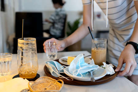 Hand of female waitress holding a tray with scraps of food,leftover food,dirty dishes,tissues and used protective face masks left on plates,girl cleaning the table in a restaurant,concept of litteringの写真素材