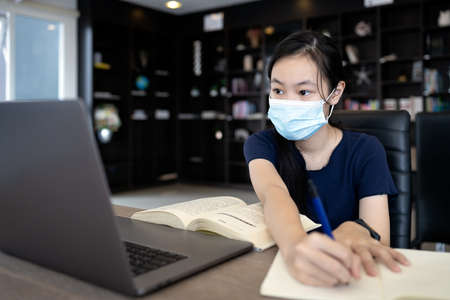 Asian student wearing protective mask,taking notes from laptop computer and textbooks in library at school,writing do homework,concentrated child girl make notes in the book prepare for test or examの写真素材