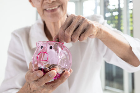 Hand of senior woman putting coins into piggy bank,Smiling elderly holding a piggy bank pink pig feel happy,saving money for health care,medical insurance,concept of financial planning in retirementの写真素材