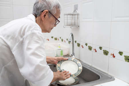 Senior woman or grandmother washing the dishes by hands in the sink,asian elderly clean dirty plates,wash dishes with dishwashing liquid and sponge under running water in a kitchen sink at homeの写真素材
