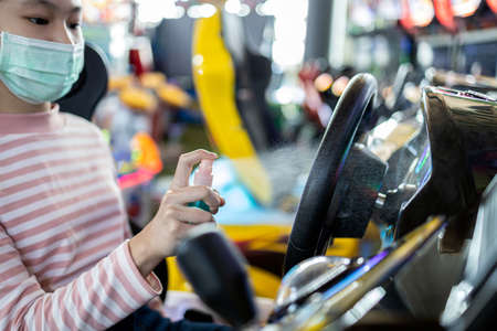 Child girl in protective mask,kid girl spraying alcohol disinfectant,clean,hygienic,sanitizing dirt,prevent infection  virus on steering wheel of toy car racing game before playingの写真素材