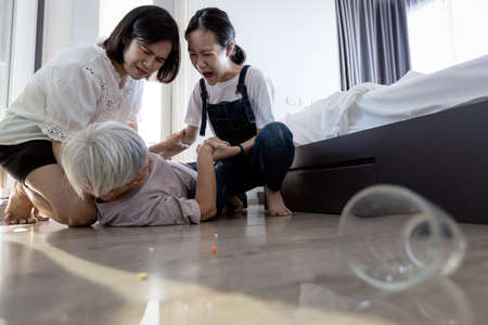 Shocked daughter and granddaughter helping to support her old mother fell unconscious lying on the floor,lift her up to stand up after falling down,asian elderly people in a coma from slipping at homeの写真素材