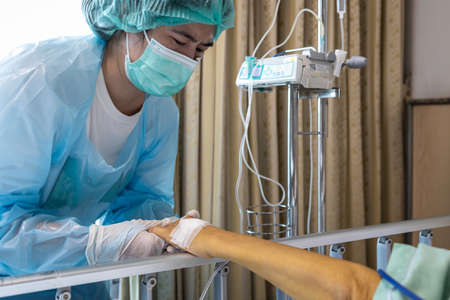 Asian daughter in protective clothing,wear medical face mask and gloves,woman holding hands,visit her mother who was suffering from a contagious disease,taking care of elderly in a coma at hospitalの写真素材