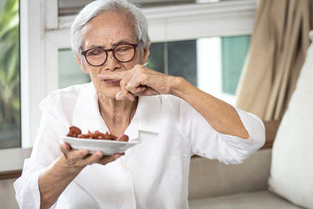Senior woman holding plate of bad spoiled or expired food in her hand,rotten food,emitting a fetid smell or strong-smelling food,disgusted old elderly cover nose with her finger,diet,nutrition conceptの写真素材