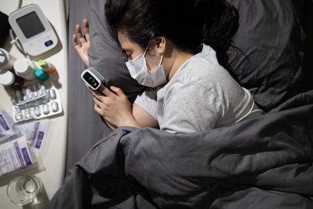 Infected asian woman lies in bed,taking care of herself with medication from doctor,fever from virus infection during pandemic,quarantine,self-isolation,medical treatment at her homeの写真素材
