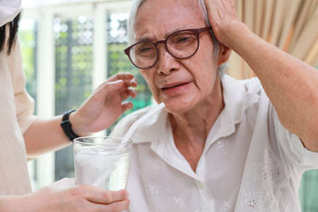 Closeup,glass of water with ice,senior grandmother with a brain freeze after drinking cold water,severe pain in her head,Asian elderly woman getting brain freeze,health care,food and drinks conceptの写真素材