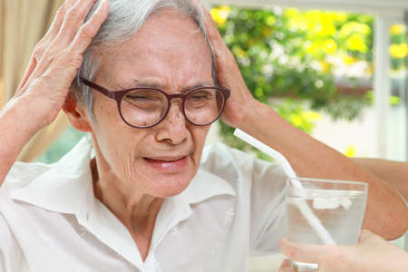 Thirsty senior woman with a brain freeze after drinking cold water with ice,Asian old elderly touching her head with hands,pain in head induced by the consumption of very cold drink,drink it too fastの写真素材