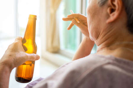 Lonely old elderly people with alcoholic drink and cigarette in her hand,sad depressed senior woman drinking and smoking while alone at home suffering from alcoholism,Addiction and depression conceptの写真素材