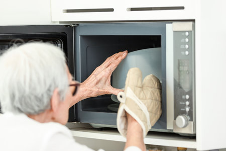 Asian senior woman wearing oven glove while taking food out of microwave oven,prevent damage and injury from heat accident,safety at home,hot bowl from cooking,old elderly heating food in kitchenの写真素材