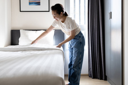 Asian young woman making bed arranging blanket,changing bedding in her room,happy smiling teen girl cleaning,tidying up the bedroom,enjoy while doing housekeeping at home,housework, hygiene conceptの写真素材