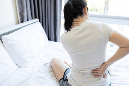 Asian adult woman have pain in neck,shoulder and backache sitting on bed in bedroom,Stressed people suffering from back pain,tension and injury in her nape of neck on uncomfortable pillow and mattressの写真素材