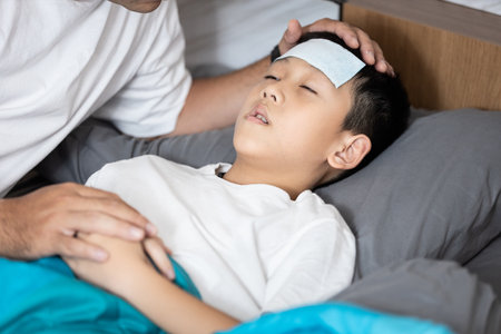 Child boy suffering from flu,influenza virus,fever and cold,kid has cooling gel on forehead,rest and sleep on bed at home,Loving caring father touching head,holding hand,take care,look after sick sonの写真素材