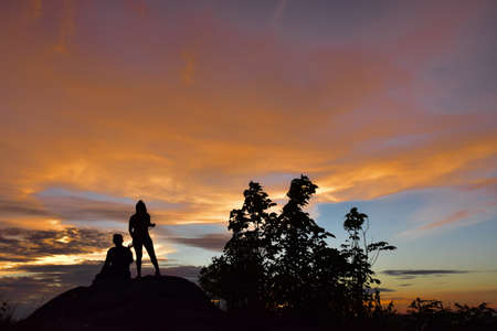 Men and women sit and watch the sunset on a high mountain peak with a glowing yellow sky. Silhouette photography techniques.の写真素材