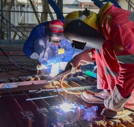 Two welder is assembling the workpiece by process shielded metal arc welding (SMAW). Welder in red uniform, safety shoes, leather gloves, welding mask. He is sitting and welding.の写真素材