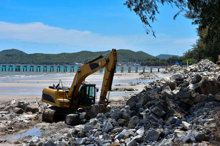 Crawler excavator are moving rocks to make breakwater on the beach. The beach , mountains and blue sky blackgroundの写真素材