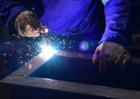 Welder is assembling the workpiece by process shielded metal arc welding (SMAW). Welder in blue uniform, safety shoes, leather gloves, welding mask. He is sitting and welding.の写真素材