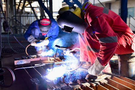 Two welder is assembling the workpiece by process shielded metal arc welding (SMAW). Welder in red uniform, safety shoes, leather gloves, welding mask. He is sitting and welding.の写真素材