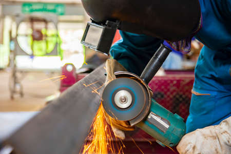 worker in blue uniform cutting metal sheets with electric grinder in the workshop. Mechanic wear personal protective equipment(PPE) while working.Leather gloves and face shield.の写真素材