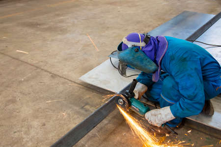 worker in blue uniform cutting metal sheets with electric grinder in the workshop. Mechanic wear personal protective equipment(PPE) while working.Leather gloves and face shield.の写真素材