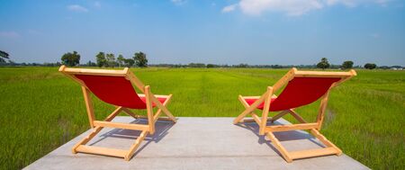 The back view of Two wooden folding chairs For sitting on the lost area The backseat is red and the seat is empty. The background is lush green rice fields and the sky is beautiful, with some clouds.の写真素材