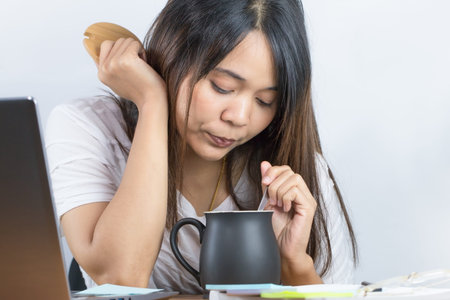 Asian woman wearing a white T-shirt Working at home Looking down at the black coffee mug on the desk With a laptop computer and a white wall backgroundの写真素材