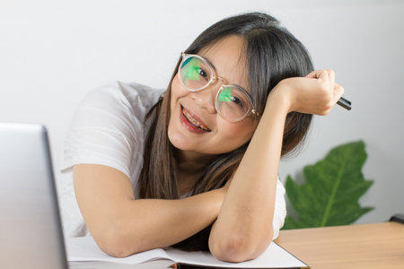 Asian woman wearing a white T-shirt and wearing glasses to work at home with happy emotions Eyes looking at the screen of a laptop computer Placed on desk and hand holding a pen and white backgroundの写真素材
