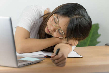 Asian woman wearing a white T-shirt and wearing glasses to work at home with happy emotions Eyes looking at the screen of a laptop computer Placed on desk and hand holding a pen and white backgroundの写真素材