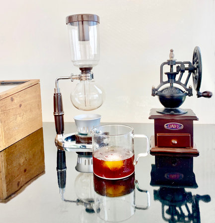 Japanese siphon vacuum coffee maker, black coffee in a clear coffee mug, coffee grinder, and coffee bean storage wooden box. all placed on a clear, reflective glass table and a window curtain as a background.の写真素材