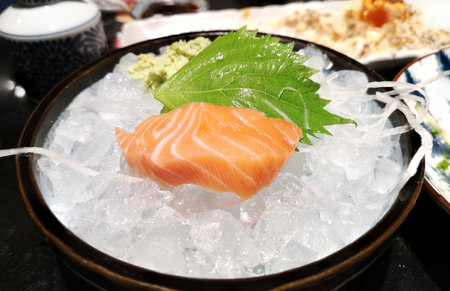 Salmon sashimi cutting fresh raw piece is a Japanese delicacy with green oba leaf and wasabi on ice in black bowl.の写真素材