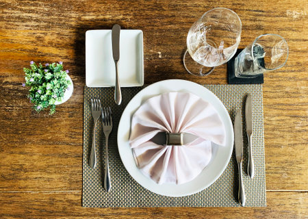 Flat lay horizontal photo of Italian dining table arrangement. A clean utensil with knife, fork, white fabric napkin, wine and water glasses on a cotton mat and wooden table, and a flowerpot.の写真素材