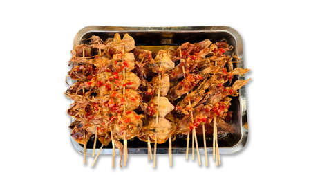 Close-up horizontal image of grilled dried squid on skewers topped with dipping sauce in the stainless tray isolated on the white background. One of Thailand's most famous street food, the concept of Asian traditional appetizer. Top-view macro photography of dried cuttlefish BBQ roasted skewer grill topped with spicy dipping sauce.の写真素材