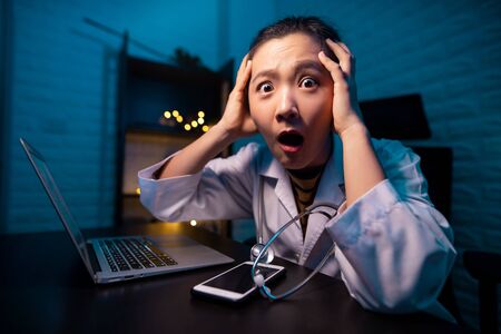 Asian woman doctor panic working and video call on a laptop overtime at home. WFH. Work from home for avoid the Coronavirus COVID 19 concept.の写真素材