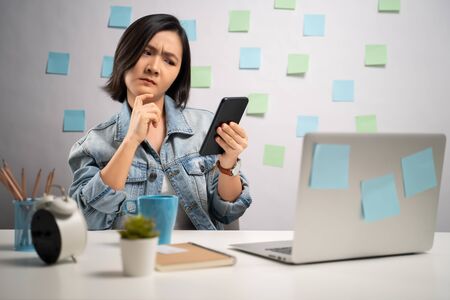 Asian woman looking at smart phone reading news and feel confused at home office. WFH. Work from home. Prevention Coronavirus COVID-19 concept.の写真素材