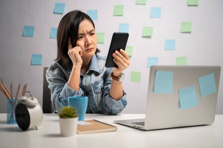 Asian woman looking at smart phone reading news and feel confused at home office. WFH. Work from home. Prevention Coronavirus COVID-19 concept.の写真素材