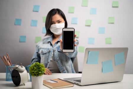 Asian woman wearing protective face mask showing blank screen at home office. WFH. Work from home. Prevention Coronavirus COVID-19 concept.の写真素材