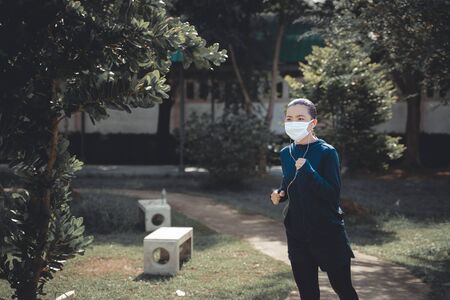 Portrait of Asian woman running at village park listening to music and wearing mask. Woman wearing mask for protective pollution and protection virus, coronavirus and running at park.の写真素材