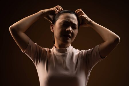 Asian woman bored and annoyed, scratching her head, standing isolated on beige background. Low key.の写真素材