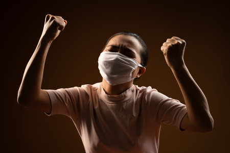 Asian woman wearing protective face mask showing her fist make a winning gesture standing isolated on beige background. Low key.の写真素材