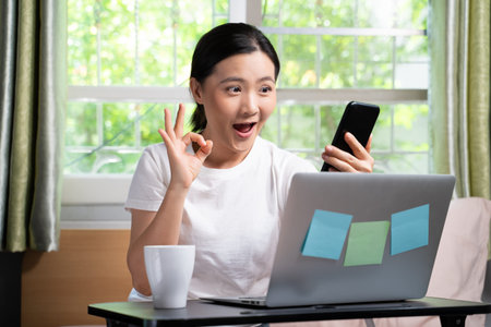 Asian woman happy video call with laptop holding smart phone sitting on the bed and showing OK sign...