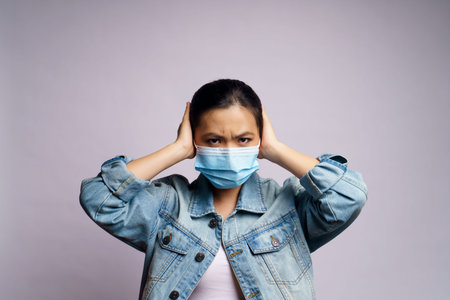 Asian woman wearing protective face mask and covering her ears by hands isolated on white...