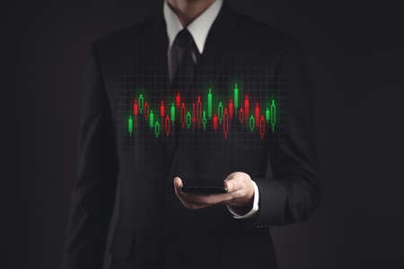 Businessman in black suit using smartphone planning for investment. Showing candlestick chart hologram technology.の写真素材