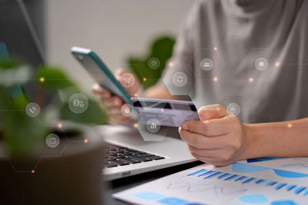 Close up shot of females hands holding credit card typing message on smart phone with technology icons for shopping online at home office.の写真素材