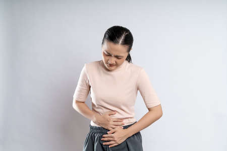 Asian woman was sick with stomach ache, putting her hands on her belly and standing isolated on white background.の素材 [FY310190264285]