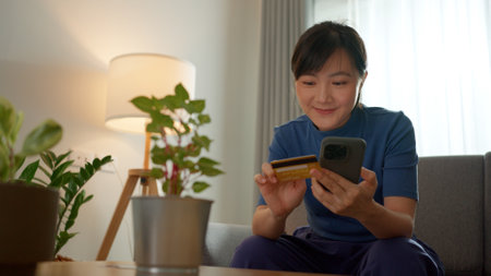 Asian woman sitting on sofa using smart phone and credit card for shopping online at home. Happy woman shopper using instant mobile payments making purchase in online store.の写真素材