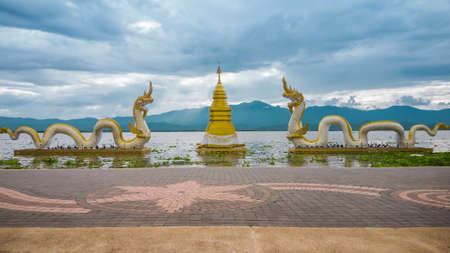 Golden pagoda surrounded by 2 figures of naga with the sky, white cloud, sunlight and mountain in Phayao lake (Kwan Phayao) as background. This place is located in Phayao in the northern of Thailand.の写真素材