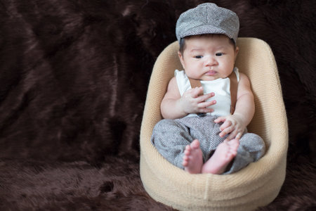 A newborn boy wearing a casual white shirt and hat with gray pants is sitting on a baby sofa on a brown background.の写真素材