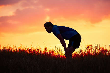 Silhouette of runner. Outdoor cross-country running. Pensive young man is taking rest after running in the nature during golden sunset.の写真素材