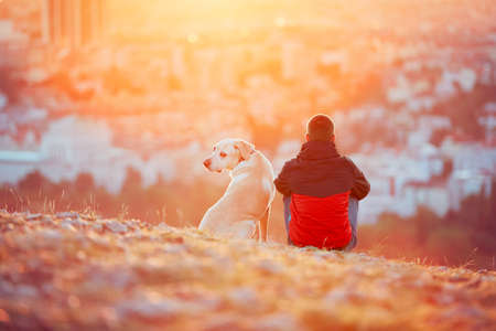Enjoying sun. Pensive young man sitting on the hill with his dog (yellow labrador retriever). Amazing sunrise in the city. Prague in Czech Republic.の写真素材