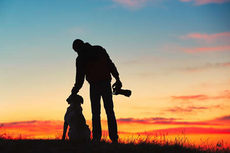 Silhouette of young photographer with his dog (labrador retriever) is enjoying sun. Photographer with mirror camera is ready to take sunrise pictures.の写真素材