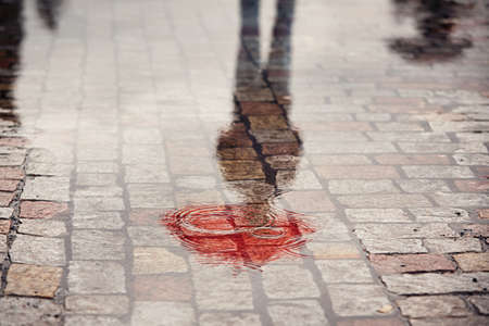 Rainy day. Reflection of young man with red umbrella in puddle on the city street during rain.の写真素材